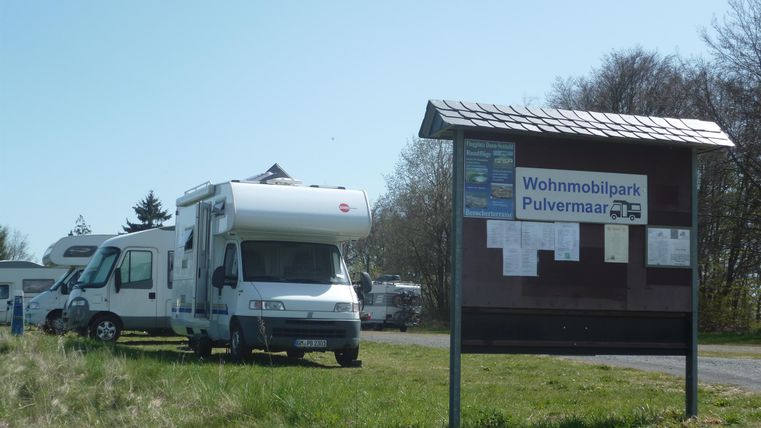 Ein Wohnmobil steht auf einem Campingplatz neben einem Informationsschild. Die Umgebung ist ruhig und grün, ideal für einen Urlaub im Freien.