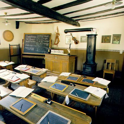 Ein traditionelles Klassenzimmer mit Holztischen und einer Tafel. Die Wände sind mit Lehrmaterial und Bildern dekoriert.
