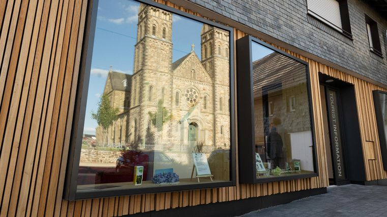 Ein modernes Gebäude mit Holzelementen und großen Fenstern spiegelt eine wunderschöne Kirche wider. Die Kirche hat zwei Türme und ist von Bäumen umgeben.