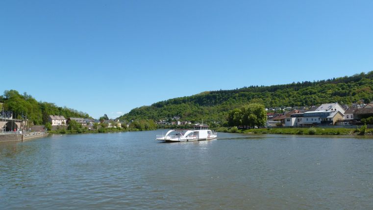 A calm river with a boat on the water. The banks are surrounded by green hills and residential houses.