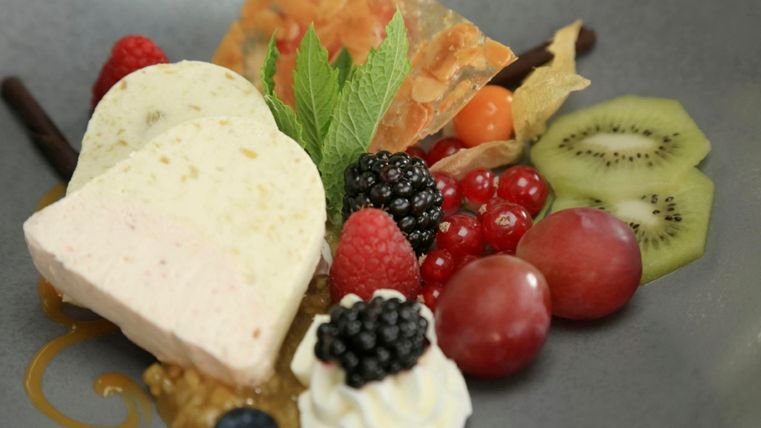 An artistically arranged dessert with a heart shape made of ice cream, fresh berries, and fruity garnishes. Green fruits and a decorative sweet treat complement the dish.