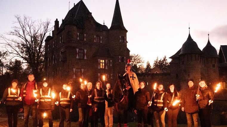 A group of people stands with torches in front of an old castle. In the background, the sky is in twilight and the architecture of the castle is visible.
