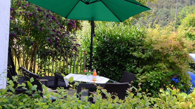 A cozy seating area in the garden with a green sun umbrella. The table is set for a small refreshment, surrounded by lush greenery.