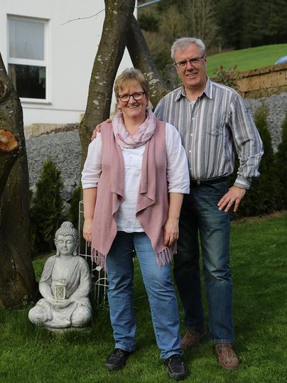 Ein Paar steht im Garten neben einer Buddha-Statue. Im Hintergrund sieht man ein Haus und viel Grün.