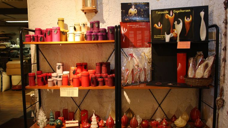 Ein Regal mit verschiedenen Kerzen in verschiedenen Formen und Farben. Auf dem Regal sind auch einige dekorative Figuren und Plakate zu sehen.