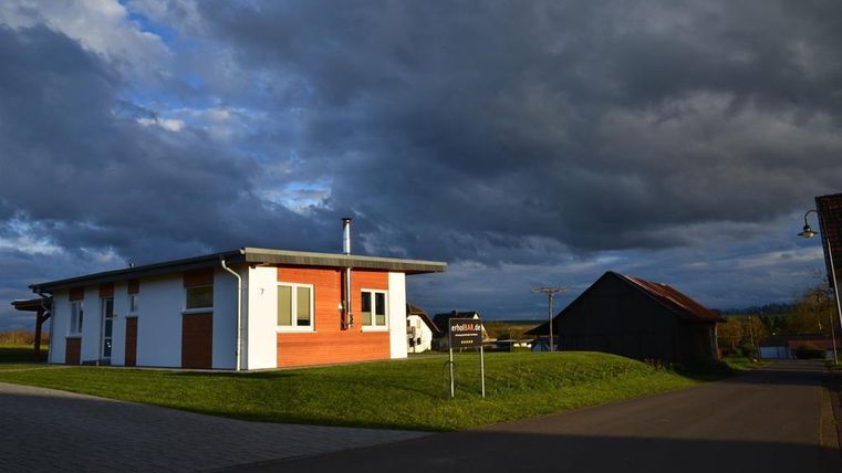 A modern house with a red facade stands in a rural setting. Dark clouds are passing by in the sky.