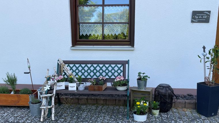 Een gezellige bank voor een wit huis, omringd door verschillende planten en bloemen. Het uitzicht vanuit het raam toont een groene landschap op de achtergrond.