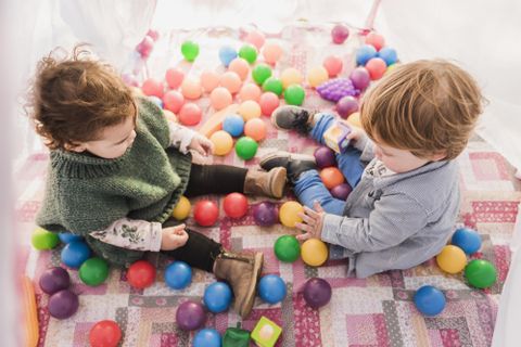 Twee kleine kinderen spelen in een kleurrijk ballenbad. Ze zitten op een deken, omringd door veel kleurrijke ballen.
