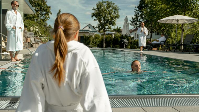Eine entspannte Szene am Pool mit mehreren Personen in Bademänteln. Zwei Personen stehen am Rand, während eine andere im Wasser schwimmt.