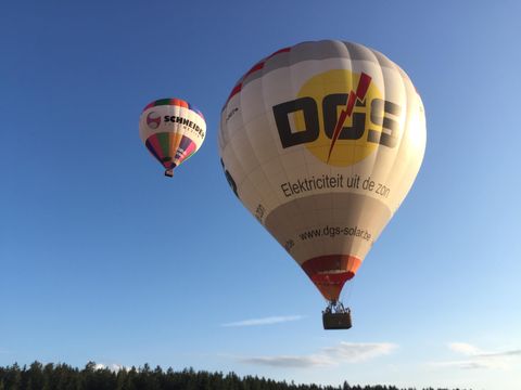 Twee luchtballonnen zweven boven een boslandschap. De grotere ballon heeft het logo van een bedrijf en straalt in de avondzon.