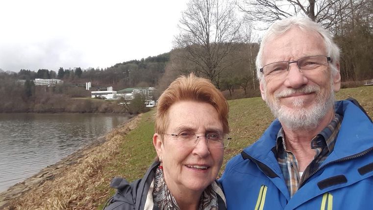 Ein Paar lächelt am Ufer eines Sees. Im Hintergrund sind Bäume und eine hügelige Landschaft zu sehen.