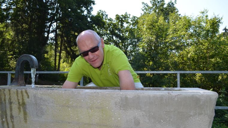 Een man in een lichtgroen shirt leunt over een betonnen rand. Op de achtergrond zijn groene bomen en een blauwe lucht te zien.