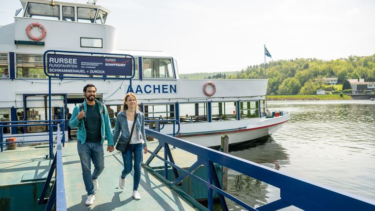A group of people walks along a dock. In the background, a tour boat is visible on the water.