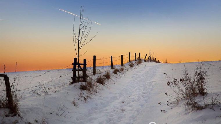 Een besneeuwd pad leidt naar een hek tegen een oranje zonsondergang. Aan de hemel zijn lichte wolken en een vliegtuigtrein te zien.