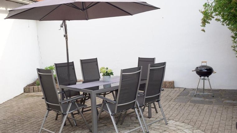 Ein gemütlicher Außenbereich mit einem Esstisch und sechs Stühlen unter einem Sonnenschirm. Neben dem Tisch steht ein Grill auf einem gepflasterten Boden.