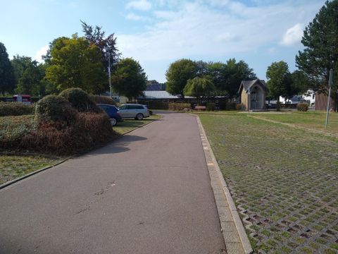 Eine ruhige Straße mit parkenden Autos und üppigem Grün. Im Hintergrund sieht man ein kleines Gebäude und Bäume.