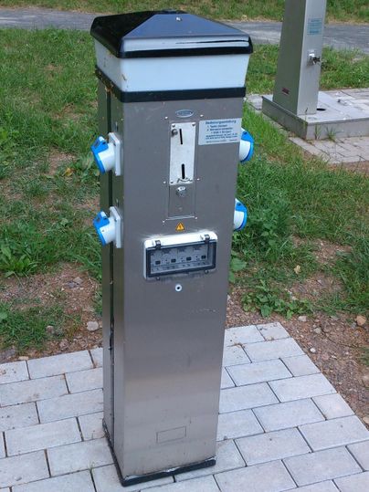 Eine moderne Säule zur Stromversorgung mit mehreren Anschlüssen. Sie steht auf einem gepflasterten Bereich in einer grünen Umgebung.