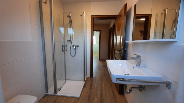 A modern bathroom with a shower and a sink. The walls are bright, and the floor is designed with wood effect tiles.