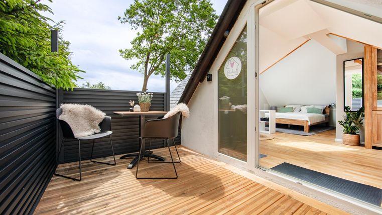 Een modern terras met houten vloer en twee stoelen. Op de achtergrond is een lichte kamer zichtbaar met een bed en planten.