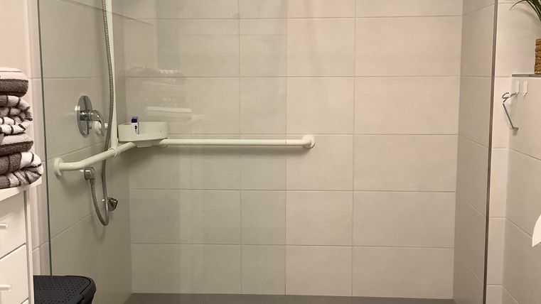 Ein modernes Badezimmer mit einer barrierefreien Dusche und einem WC. Die Wände sind hell gefliest, und es gibt Handläufe für zusätzliche Sicherheit.