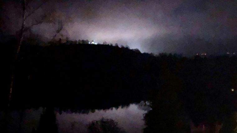 Een donkere nachtlanschap met een rustige meer en lichte nevel. Op de achtergrond zijn zwakke lichten te zien.