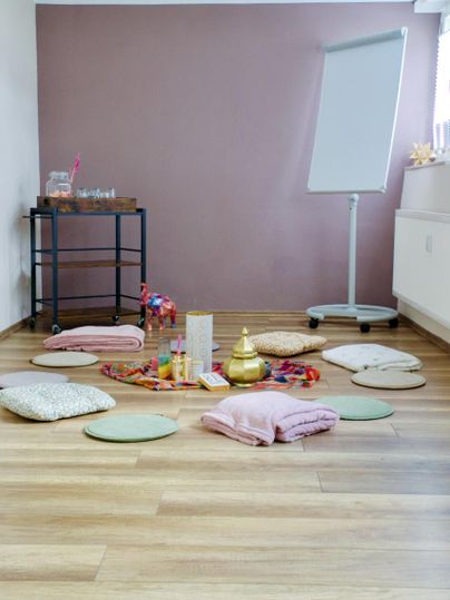 Ein einladender Raum mit bunten Kissen und Dekoration auf dem Boden. Im Hintergrund steht ein Flipchart und ein Tisch mit weiteren Utensilien.