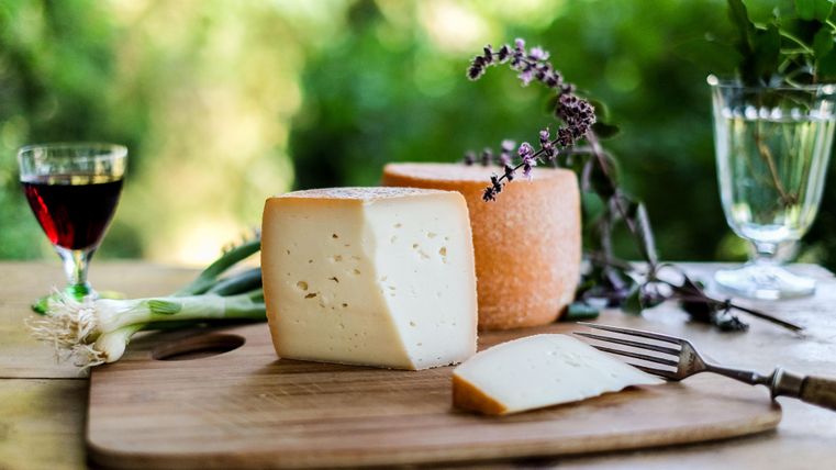 Ein Holzbrett mit zwei Käsestücken, einer halbierten und einer ganzen, umgeben von frischen Kräutern und einer Zwiebel. Im Hintergrund stehen zwei Gläser, eines mit rotem Getränk und das andere mit klarem Flüssigkeit.