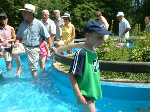 Eine Gruppe von Menschen steht in einem blauen Wasserbecken, während ein Junge im Vordergrund spielt. Die Umgebung ist grün und sonnig, was eine fröhliche Atmosphäre vermittelt.