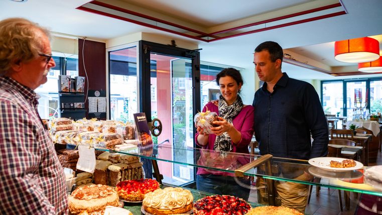 Ein Paar steht vor einer Theke mit einer Auswahl an Kuchen und Gebäck. Sie betrachten ein Dessert, während ein Verkäufer ihnen gegenübersteht.