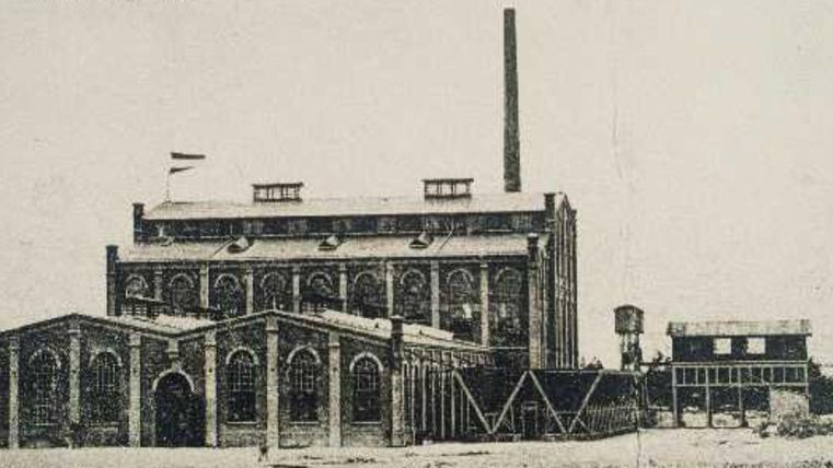 Een historisch fabrieksgebouw met een hoge schoorsteen en grote ramen. Op de voorgrond zijn extra gebouwen en een watertank zichtbaar.