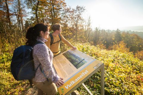 Zwei Wanderer betrachten ein Informationsschild über die Umgebung. Die Szene zeigt eine herbstliche Landschaft mit bunten Bäumen und klarem Himmel.