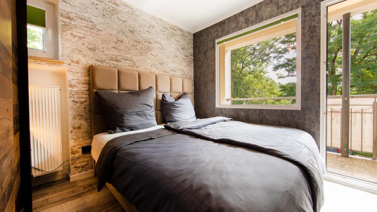 Ein modernes Schlafzimmer mit einem Doppelbett und stilvollen Kissen. Große Fenster bieten Blick auf die grüne Umgebung.