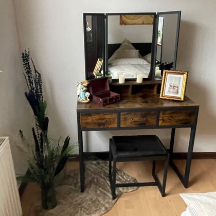 Een stijlvolle make-uptafel met een grote spiegel en een bloemdecoratie. Naast de tafel staat een kruk en een fotolijst.