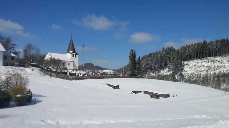 A snowy landscape with a small, white village and a church in the background. The sky is clear and blue, and trees are covered with snow.
