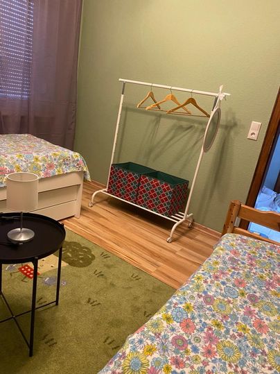 Ein einfaches Zimmer mit zwei Betten, einem Kleiderständer und einem Tisch. Die Wände sind grün und die Böden sind aus Holz.