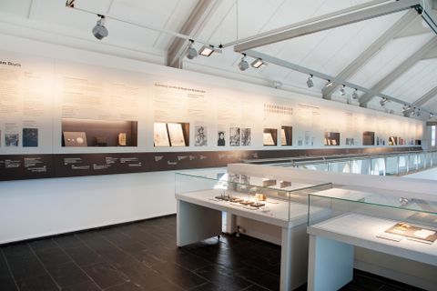 Een modern museum met een wand vol informatie en tentoonstellingen. Op de voorgrond zijn vitrines met verschillende objecten te zien.