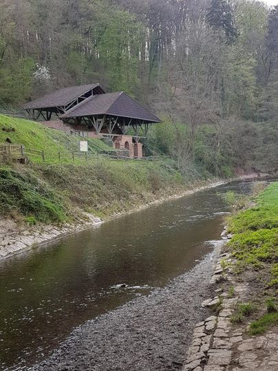 Ein ruhiger Fluss fließt entlang einer grünen Uferlandschaft. Im Hintergrund ist ein Gebäude mit einem schrägen Dach zu sehen, umgeben von Bäumen.