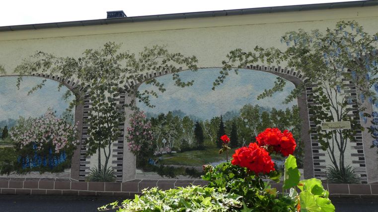 Eine farbenfrohe Wandmalerei mit Bäumen und einem Gartenblick. Im Vordergrund sind rote Blumen zu sehen.