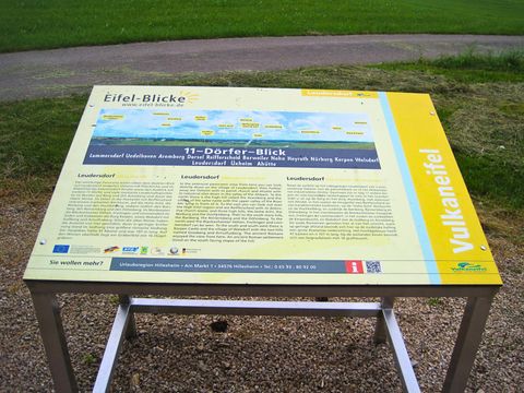 Infotafel '11-Dörfer-Blick' in der Vulkaneifel mit Informationen zu umliegenden Dörfern und Landschaften. Hintergrund zeigt Wiese und Weg.