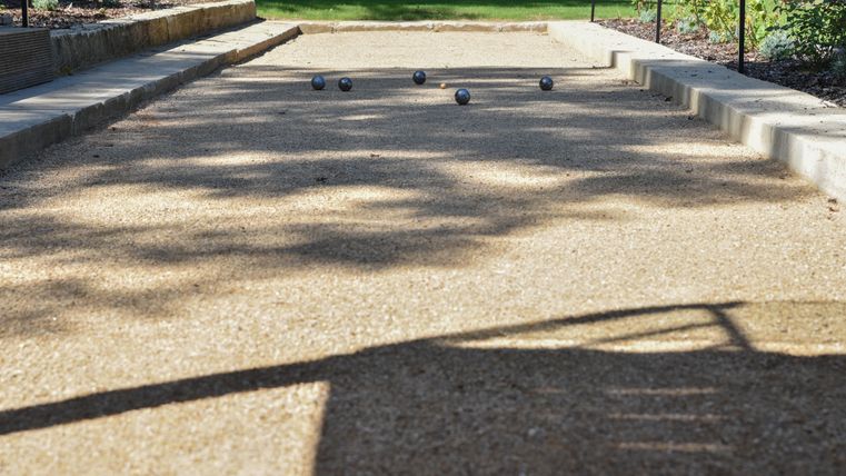 Ein Bocciabahn mit mehreren Kugeln auf dem Kiesboden. Im Hintergrund sind grüne Pflanzen und ein sonniger Garten zu sehen.