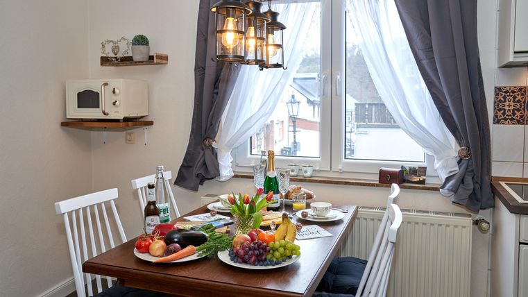 A cozy dining area with a wooden table and white chairs. Various foods and drinks are arranged on the table, while the light falls through the windows.