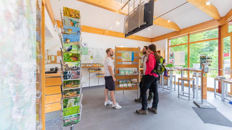 Ein helles Informationszentrum mit großen Fenstern und Holzdetails. Drei Personen schauen sich Karten und Informationen an.