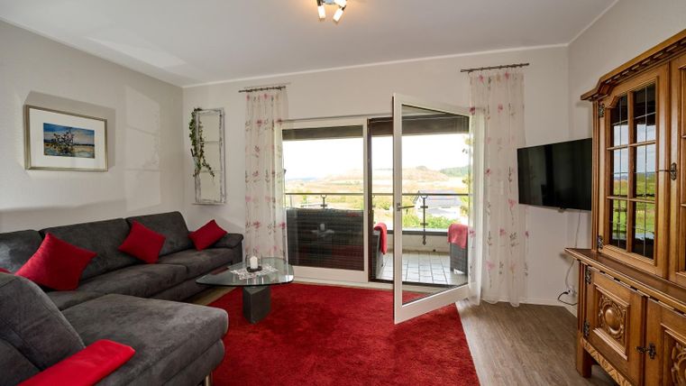 Een moderne woonkamer met een grijze bank en rode kussens. Grote ramen bieden uitzicht op het landschap en de ruimte is helder en uitnodigend.