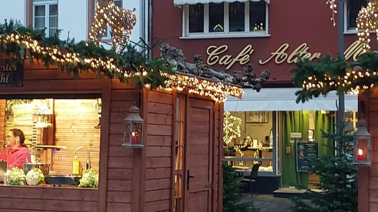Eine festlich dekorierte Gasse mit Holzbuden und Weihnachtslichtern. Im Hintergrund ist das Café Altenmarkt zu sehen.