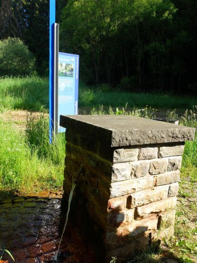 A stone fountain with flowing water in a green environment. In the background, there is an information sign.