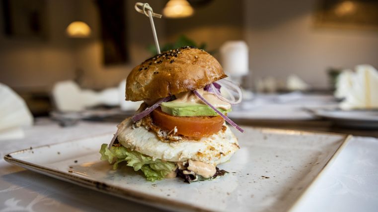Een lekkere burger op een bord, gegarneerd met sla, tomaat, avocado en uien. De burger heeft een vers broodje en wordt geserveerd in een stijlvol restaurant.