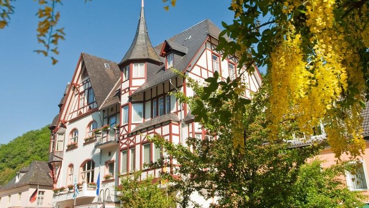 Ein schönes, historisches Gebäude mit einem spitzen Turm. Es ist von Bäumen und blühenden Pflanzen umgeben.