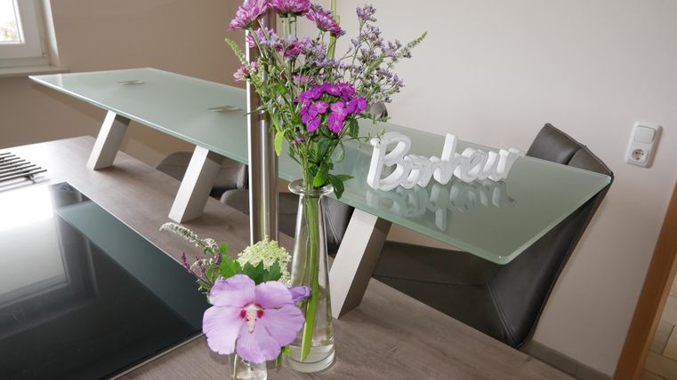 A modern dining area with a glass table and stylish chairs. Fresh flowers in vases and a decorative sign enhance the cozy atmosphere.