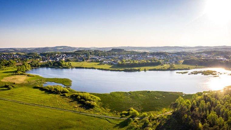Een rustige vijver omringd door groene weilanden en bossen. Op de achtergrond zijn kleine dorpen en zachte heuvels te zien.
