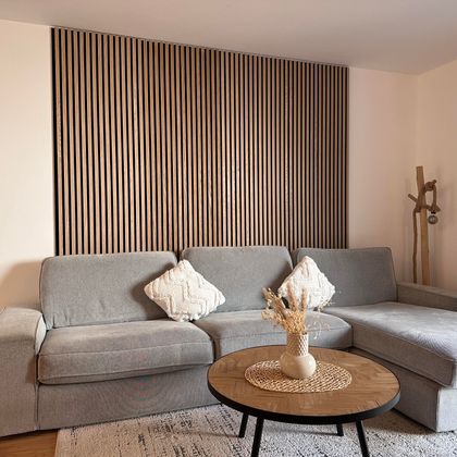 Ein modernes Wohnzimmer mit einem grauen Sofa und einem runden Tisch. Die Wand im Hintergrund ist mit Holzlamellen gestaltet.